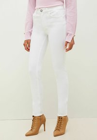 Jeans blancos ajustados con bolsillos laterales, cremallera frontal y un ligero brillo, combinados con botas de tacón alto de color marrón claro con cordones y punta afilada.