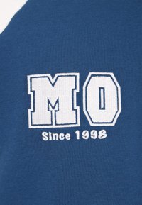 Kék szövet fehér hímzett betűkkel, "MO" és alatta "Since 1998" felirattal. A minta puha textúrájú és erőteljes, egyetemi stílusú.