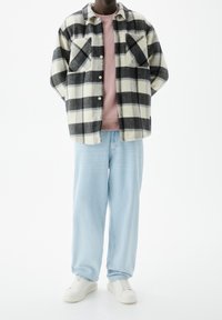 Persoon in een zwart-wit geblokte jas over een roze shirt, lichte blauwe jeans en witte sneakers, staand met de handen achter de rug.