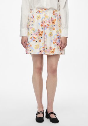 Woman wearing a white floral skirt with pastel orange, pink, yellow, and blue flowers, a white long-sleeve shirt, and black Mary Jane shoes.