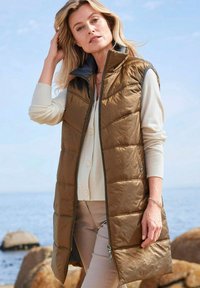 Braune wattierte Weste mit hohem Kragen, ausgestattet mit horizontalen Nähten und einem Reißverschluss, kombiniert mit einer hellbeigen Strickjacke.