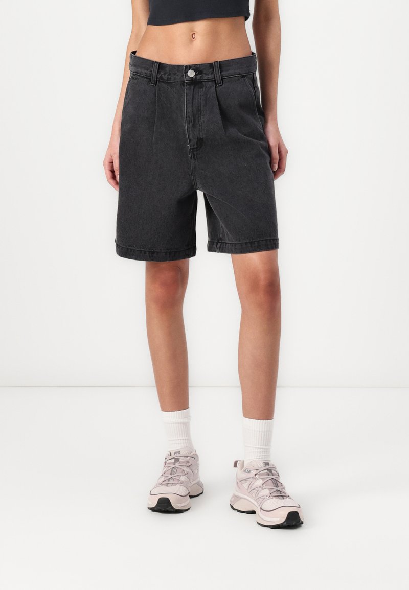 Zwarte denim shorts met een comfortabele pasvorm, met een hoge taille, plooien aan de voorkant en opgerolde hemden. Gecombineerd met lichtgekleurde sportschoenen en witte sokken.