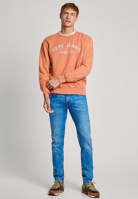 Sudadera naranja con texto blanco "PEPE JEANS LONDON W11", combinada con jeans de color azul claro y zapatillas con acentos marrones, de pie sobre un fondo neutro.