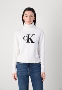 Hvit strikket turtleneck-genser med ribbede mansjetter og nederkant, med et stort svart brodert "CK"-logo på forsiden. Bært med blå jeans.