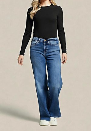 Femme portant un haut noir à manches longues, un jean flare bleu taille haute et des baskets blanches, debout devant un fond uni.