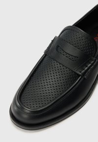 Svarte lærloafers med perforert overdel, rund tå og slip-on design. Har en rem over vristen og subtile sømdetaljer.