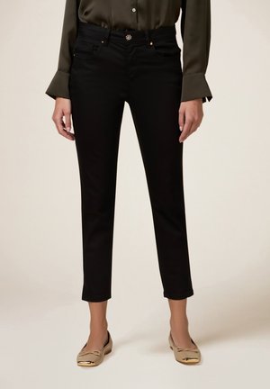 Kobieta nosząca czarne obcisłe spodnie długości do kostek, zestawione z satynową, oliwkowozieloną bluzką i beżowymi balerinami z metalowymi noskami.