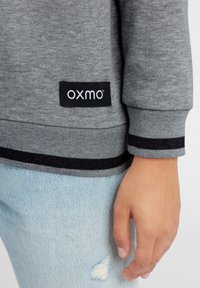 Grå hettegenser med ribbestrikkede mansjetter, med et svart logo-patch. Kombinert med lyseblå slitte denim, som viser en teksturert kontrast i stoffet.