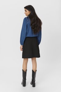 Chemisier en denim avec des manches bouffantes, associé à une jupe noire. Le mannequin porte des bottes noires jusqu'aux genoux. L'arrière-plan est blanc uni.