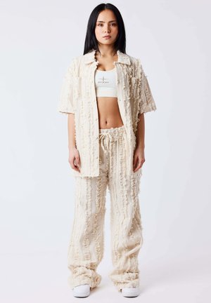 Mujer con cabello largo y negro que lleva una camisa de manga corta texturizada color crema y pantalones anchos a juego, con un top corto blanco y zapatillas blancas.