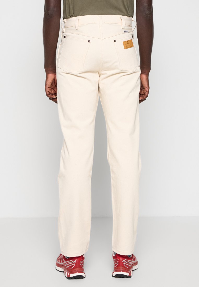 Hombre con pantalones rectos color blanco roto y zapatos deportivos rojos, de pie con las manos relajadas a los lados, vista trasera.
