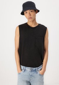 Camiseta de algodón negra sin mangas con un bolsillo frontal, combinada con unos jeans de color azul claro. La persona lleva puesto un sombrero tipo bucket negro.