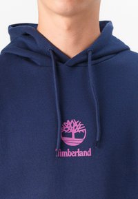 Navy blue hoodie made of soft fabric, featuring a pink Timberland logo with a tree design and drawstring hood accents.