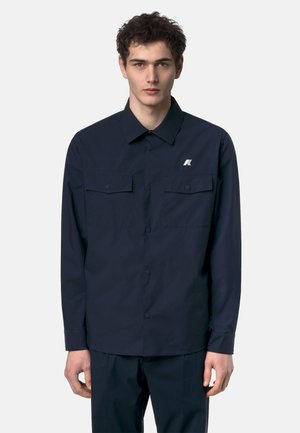 Giovane uomo con capelli corti e ricci che indossa una camicia blu navy abbottonata con due tasche sul petto e un piccolo logo bianco sul lato sinistro.