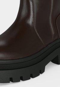 Botte en cuir marron avec semelle épaisse noire à crampons et fermeture éclair latérale visible sur fond blanc.