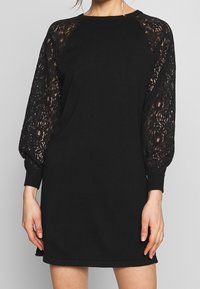 Zwarte jurk met een effen gebreide body en doorzichtige kanten pofmouwen met geribbelde manchetten. De silhouet is ontspannen en boven de knie.