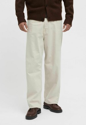 Brown knitted cardigan over a dark shirt, paired with light-colored, loose-fitting trousers and dark brown shoes with a chunky sole.