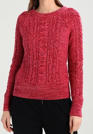 Kobieta w dopasowanym, różowym, długim rękawie swetrze z warkoczowym splotem i okrągłym dekoltem oraz czarnych spodniach na jednolitym tle.