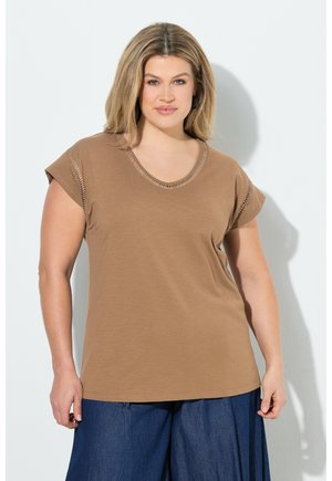 Femme aux cheveux blonds mi-longs portant un haut marron à manches courtes avec des coutures décoratives et un pantalon large bleu foncé, debout contre un mur blanc.