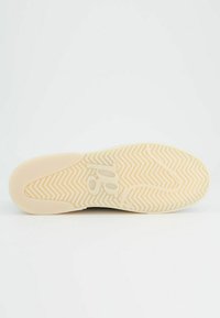 La suola di una sneaker presenta un colore crema con un motivo a spina di pesce e un logo prominente al centro. Superficie testurizzata per una buona presa.