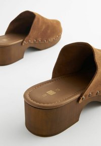 Bruine suède clogs met een houten platform, voorzien van decoratieve metalen studs langs de rand en een gladde binnenvoering.