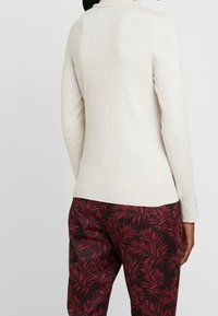 Světle béžový přiléhavý svetr s vertikálním žebrovaným vzorem, vysokým límcem a dlouhými rukávy, doplněný tmavými kalhotami s motivem červených listů.