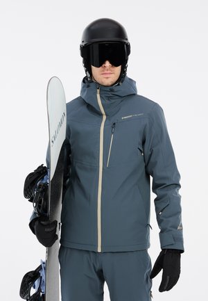 Blauwe waterdichte skijas met een hoge kraag, ritszakken en contrasterende crèmekleurige ritsen, vastgehouden door een persoon die een zwarte sk helmet en handschoenen draagt.