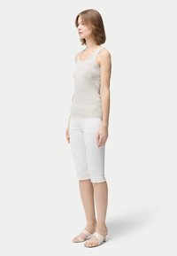 Kvinna som står i profil och bär ett ljusfärgat ärmlöst linne, vita knälånga byxor och beigea sandaler med låg klack mot en enfärgad bakgrund.