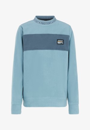 Personne portant un pull en polaire bleu avec une bande bleu foncé et un pantalon gris, debout devant un fond blanc.