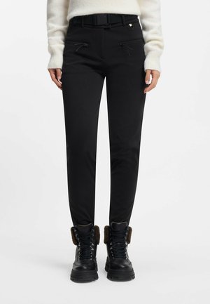 Kobieta ubrana w czarne, dopasowane spodnie z kieszeniami na zamki, czarny pasek, biały sweter i czarne sznurowane buty z brązowymi futrzanymi mankietami.