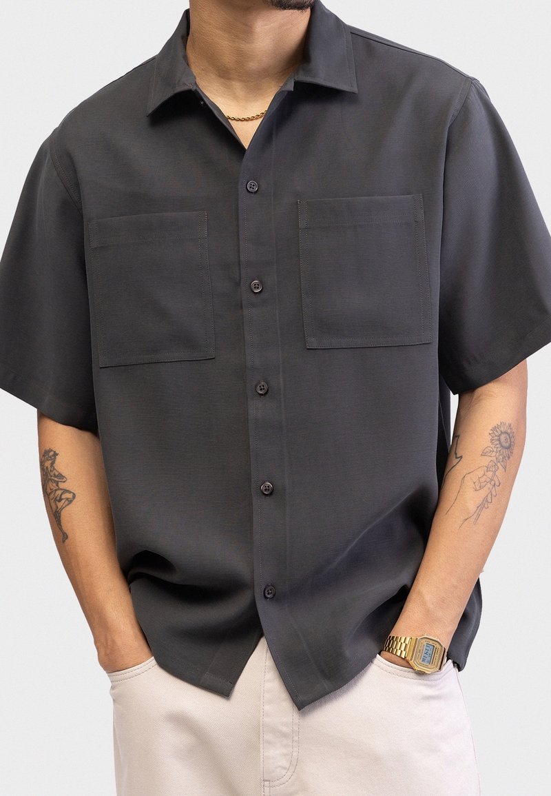 Homme portant une chemise boutonnée à manches courtes gris foncé avec deux poches sur la poitrine, pantalon beige clair, montre en or, chaîne autour du cou et tatouages sur les bras.