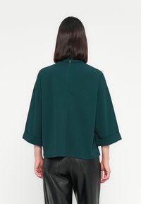 Maglia oversize in teal con colletto alto, caratterizzata da maniche ampie e una cerniera sul retro. Realizzata in un tessuto liscio, ha una vestibilità comoda.