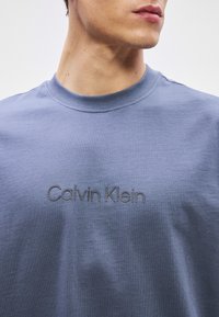 Plava pamučna majica s okruglim izrezom koja ima utisnuti "Calvin Klein" logo na prsima, glatku teksturu i opušteni kroj.
