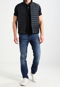 Padded black sleeveless vest with horizontal stitching, paired with a black polo shirt and blue jeans. White sneakers complete the look.