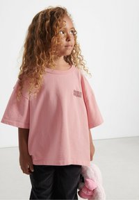 Lyserød bomulds t-shirt med løs pasform, rund halsudskæring og korte ærmer, prydet med et lille trykt logo på brystet. Bæret med mørke strukturerede bukser.