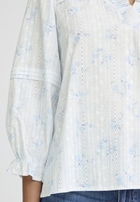 Blouse blanche à motif floral bleu, rayures verticales en œillets, manches ballon 3/4 avec poignets froncés, portée avec un jean bleu.