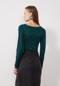 Teal long-sleeve top with ruched details, paired with a black mid-length skirt. Smooth fabric texture and fitted silhouette. Silver earrings.
