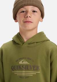 Sudadera color verde oliva con un logo gráfico frontal en marrón y beige. El diseño incluye una capucha ajustable con cordones y una textura de tela suave.
