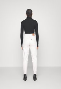 Svart långärmad topp, vita högmidjade jeans med bakfickor som har sömmar, och svarta klackade ankelstövlar. Enfärgad bakgrund.