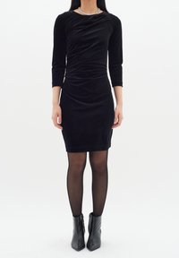 Černé sametové šaty s dlouhými rukávy, přiléhavý střih a nařasená textura. Nosí se s černými kotníkovými botami a průhlednými černými punčochami.