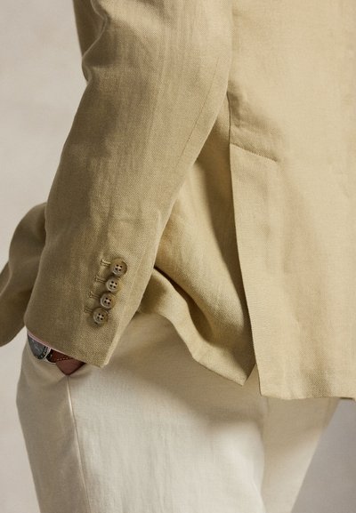 Personne portant un blazer beige avec quatre boutons sur la manche et un pantalon clair, main dans la poche, montrant le bracelet de montre.
