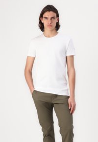 Hvid kortærmet t-shirt lavet af bomuld, med rund halsudskæring. Sammen med olivengrønne bukser, med hænderne placeret i lommerne.