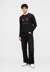 Νεαρός άνδρας που φοράει μαύρο πουκάμισο μακρυμάνικο Armani Exchange, μαύρα τζιν και λευκά αθλητικά παπούτσια, στέκεται με το ένα χέρι στην τσέπη.