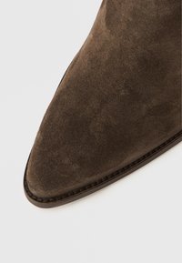 Botín de ante marrón con punta en forma de pico, con costuras mínimas y un diseño elegante. Superficie texturizada con acabado mate.