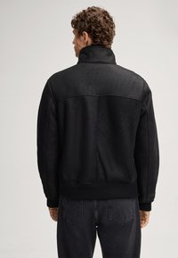 Giacca bomber nera realizzata in materiale liscio, con colletto alto e polsini e orlo a costine. Il dettaglio delle cuciture sul retro aggiunge texture.