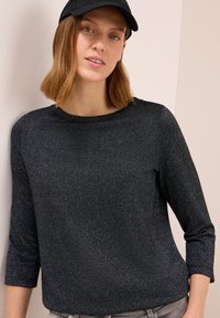 Frau mit schulterlangem Haar, die eine schwarze Mütze und ein langärmliges, dunkel glitzerndes Oberteil trägt, steht vor einer beigen Wand.