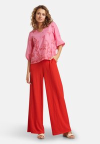 Rosa blumengemusterte Bluse mit drei Viertelärmeln, kombiniert mit roten, weit geschnittenen Textilhosen. Das Model steht in lässiger Pose.