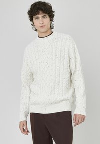 Pull en tricot blanc avec un motif en câble, col rond côtelé et finition texturée. Porté sur un pantalon marron foncé.
