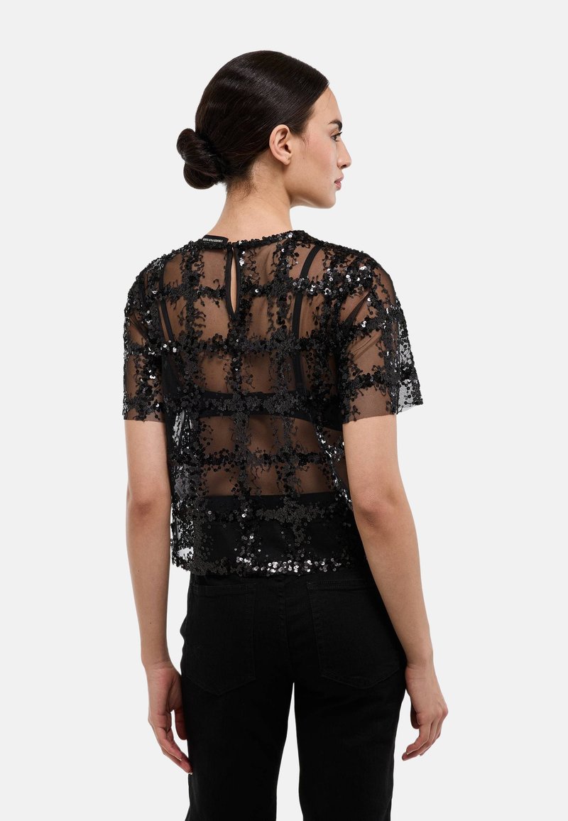 Haut noir à sequins avec une superposition en voile transparent, manches courtes et ourlet droit. Présente une ouverture en forme de goutte d'eau dans le dos. Porté avec un pantalon noir.