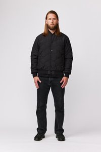 Veste bombardier matelassée noire avec un col haut, des poignets ajustés et des poches avant, associée à un jean foncé et des chaussures noires. Design simple.